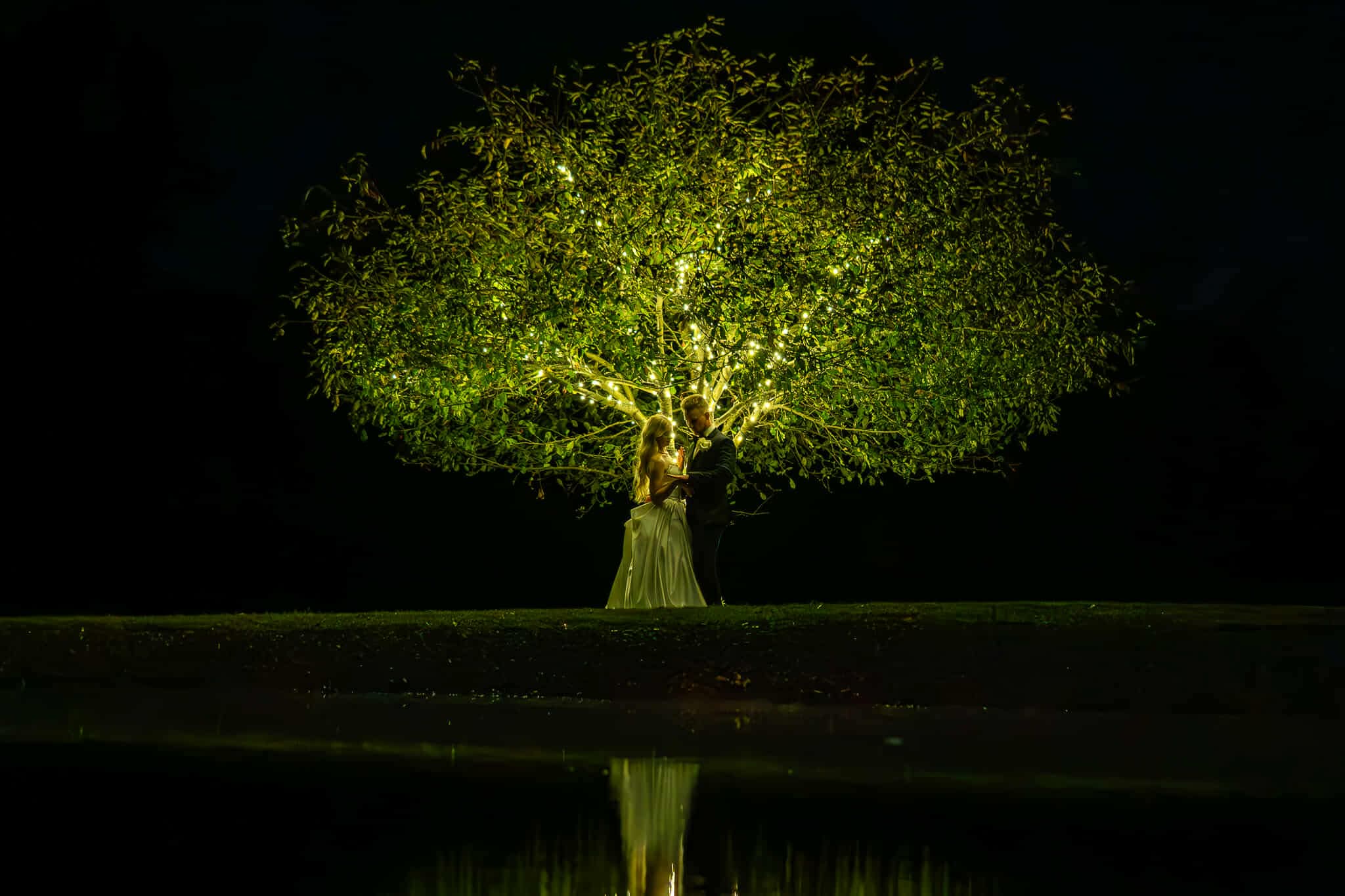Bride and groom embracing beneath a large tree wrapped in warm string lights, reflected in still water at night