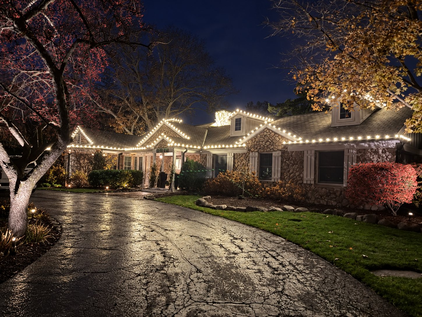 Stone rancher home with roofline lighting reflected on a wet driveway