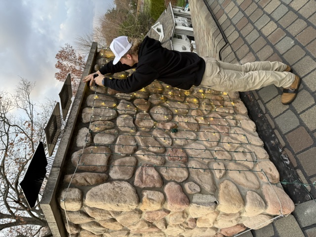 LuxeGlow technician wrapping a stone chimney in warm-white mini lights